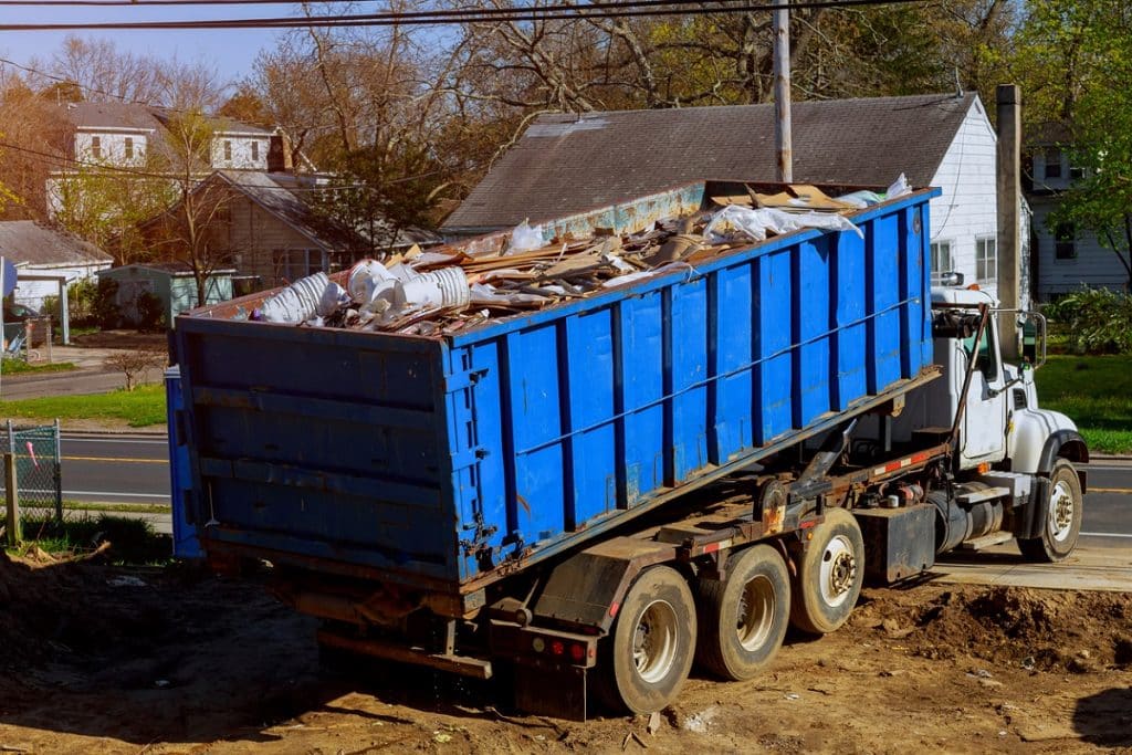 A blue dumpster truck is loaded with construction debris and household waste at a residential property.