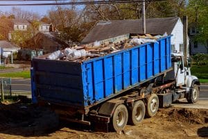 A blue dumpster truck is loaded with construction debris and household waste at a residential property.