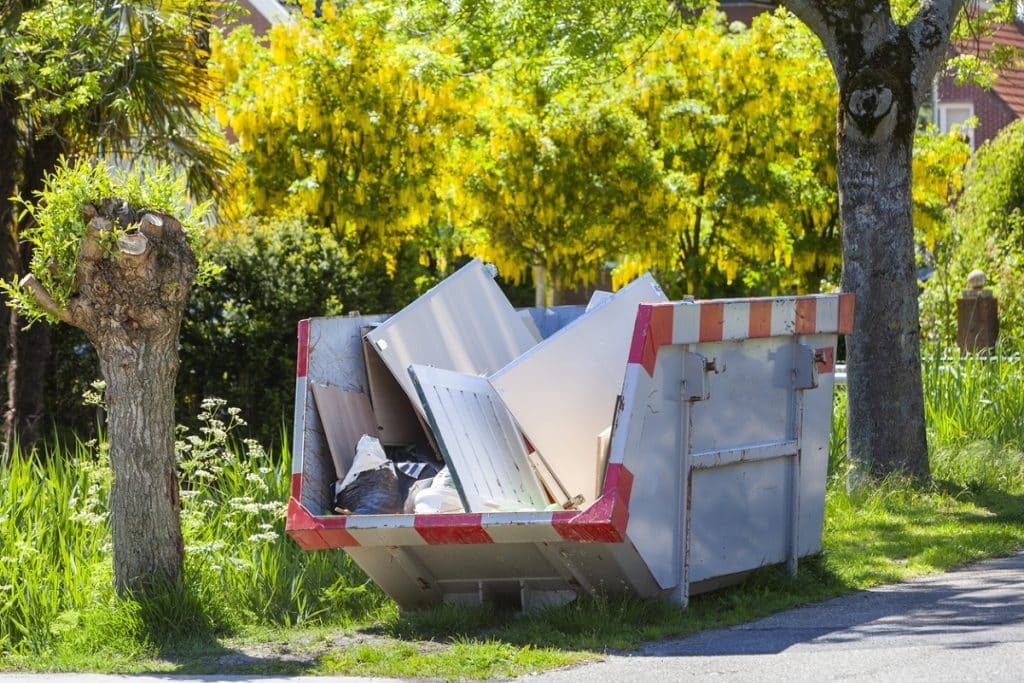 A construction dumpster filled with renovation debris, including discarded panels and building materials, beside a grassy residential property.